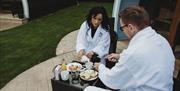 Couple having breakfast on wooden deck of glamping pod in robes