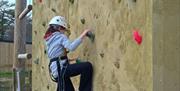 A girl in a grey jumper, blue trousers and white helmet climbing up a wall with a harness