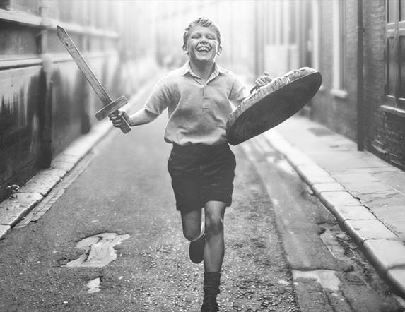 Movie poster for Belfast film, in black and white, showing a young boy running down the street with a sword and bin lid outside a row of terrace house