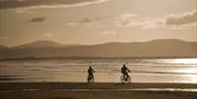 Two men on bicycles cycling down the beach with the sea and Donegal mountains in the background. Image is taken in the evening as the sun sets and the