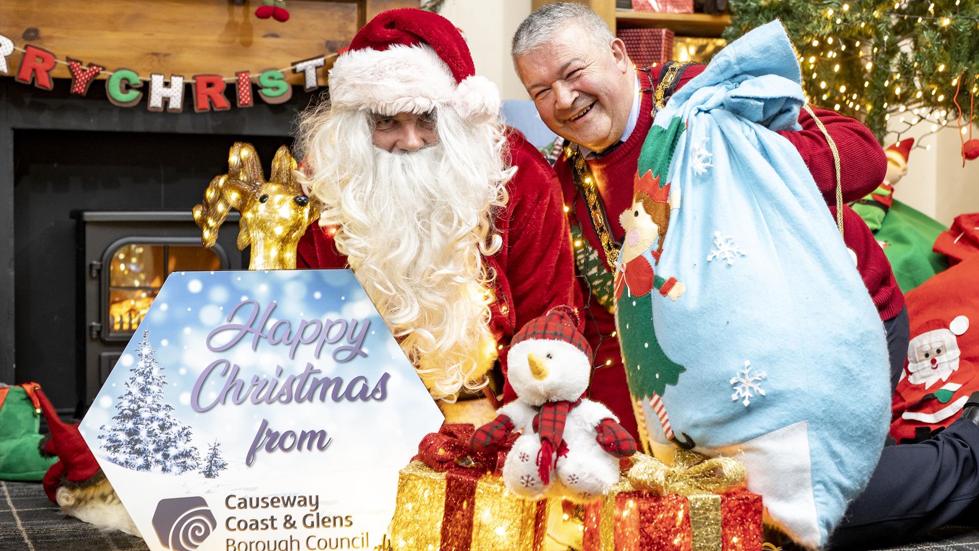 The Mayor Ivor Wallace sitting beside Santa with a blue sack, light up present and a sign reading 'Happy Christmas from Causeway Coast and Glens Borou