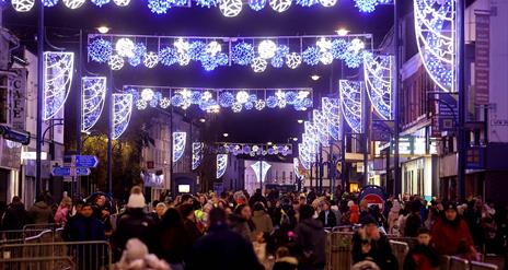 Image shows Christmas lights shining above a busy street with people shopping