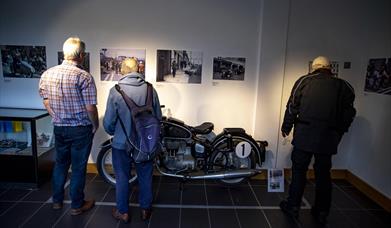 Images shows three men looking at old photographs on the wall with a vintage bike on display in front of them