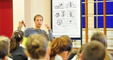 A man standing at a flip chart with a cartoon sketch on it, and the backs of children's heads watching from the audience.