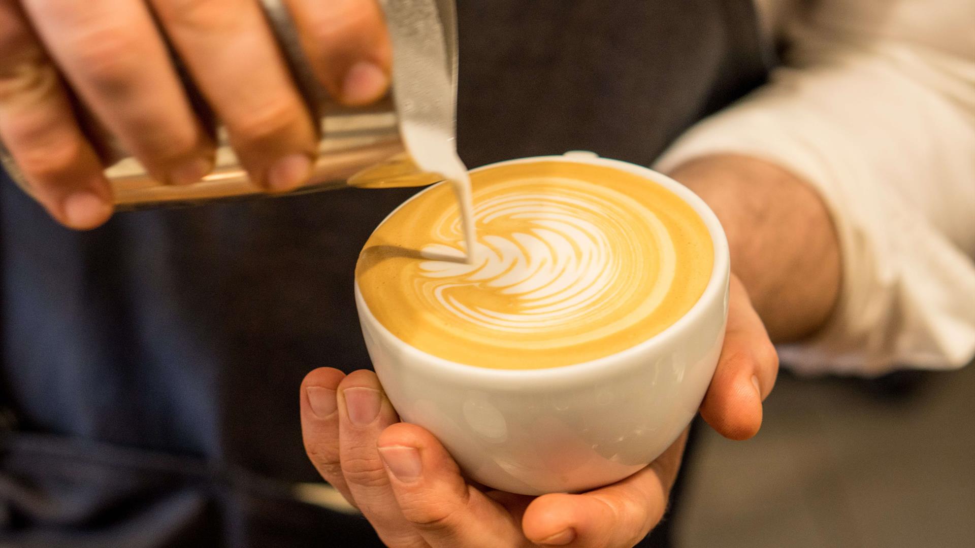 person pouring milk into latte coffee cup creating leaf pattern