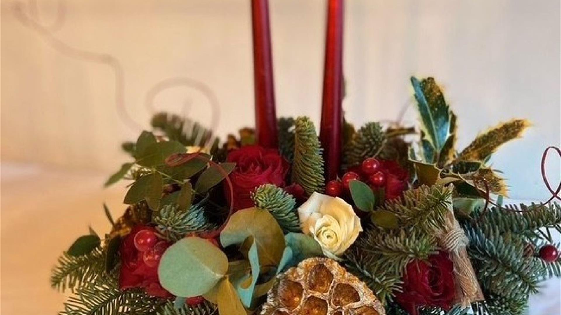 a Christmas table centrepiece decorated with holly, eucalyptus, roses and berries, with two red tapered candles coming out from the centre a Christmas table centrepiece decorated with holly, eucalyptus, roses and berries, with two red tapered candles coming out from the centre