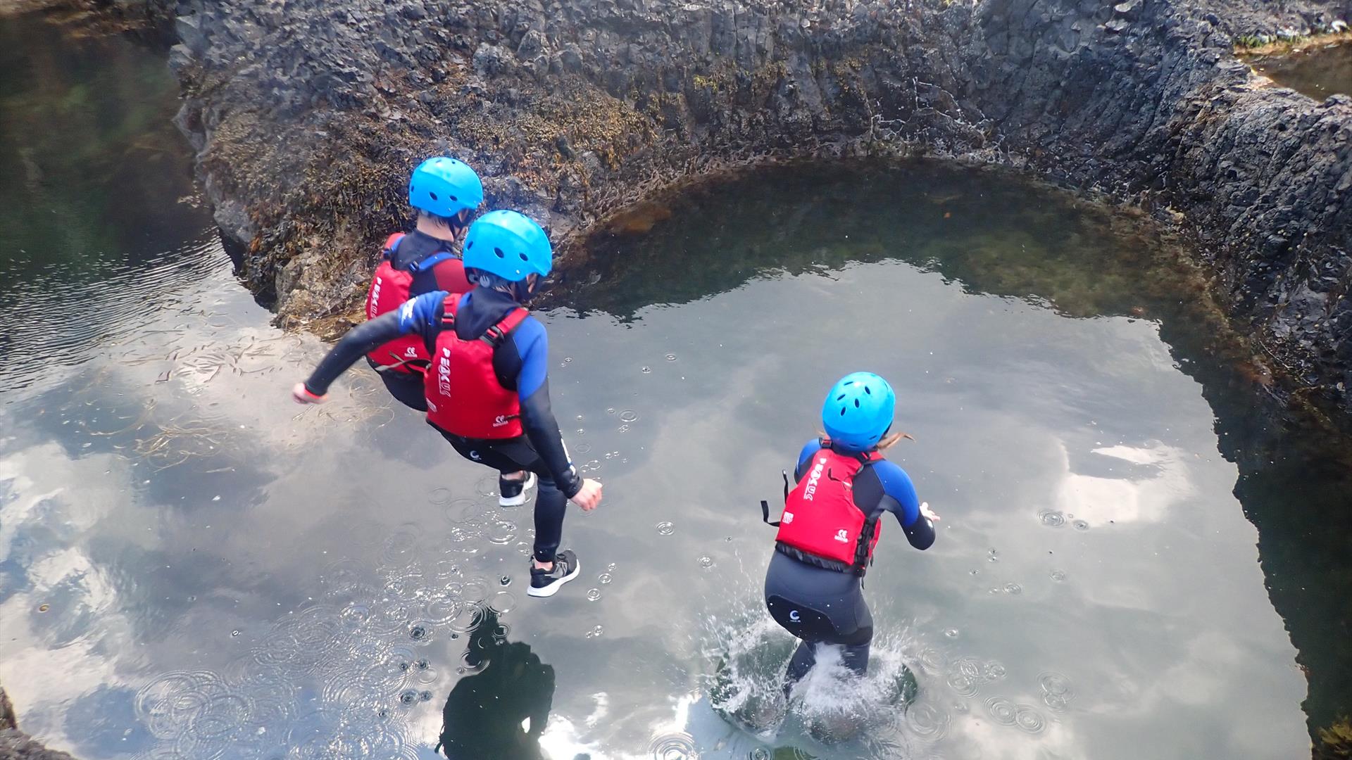 Three young children jumping into rock pool wearing helmets and buoyancy aids
