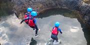 Three young children jumping into rock pool wearing helmets and buoyancy aids