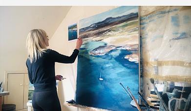 Image of Sarah Carrington artist, with blonde hair painting a canvas of  a coastal scene with art supplies in the foreground.
