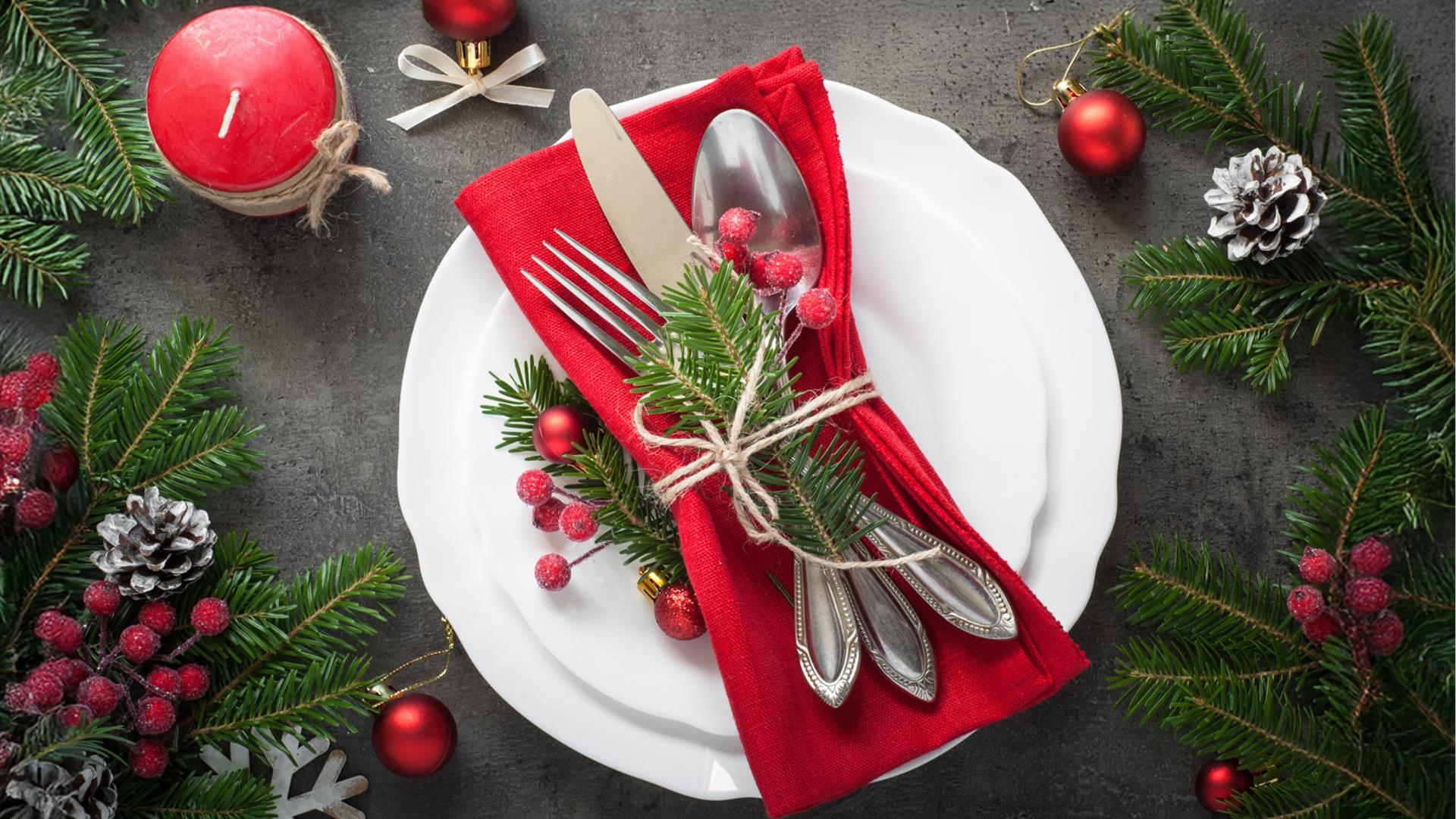 cutlery is wrapped together on a red napkin sitting on a plate, which is surrounded by festive decor including red berries, a red candle, pine cones a cutlery is wrapped together on a red napkin sitting on a plate, which is surrounded by festive decor including red berries, a red candle, pine cones a
