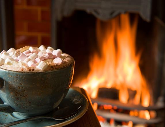 A mug of hot chocolate with marshmallows sitting in front of a roaring open fire