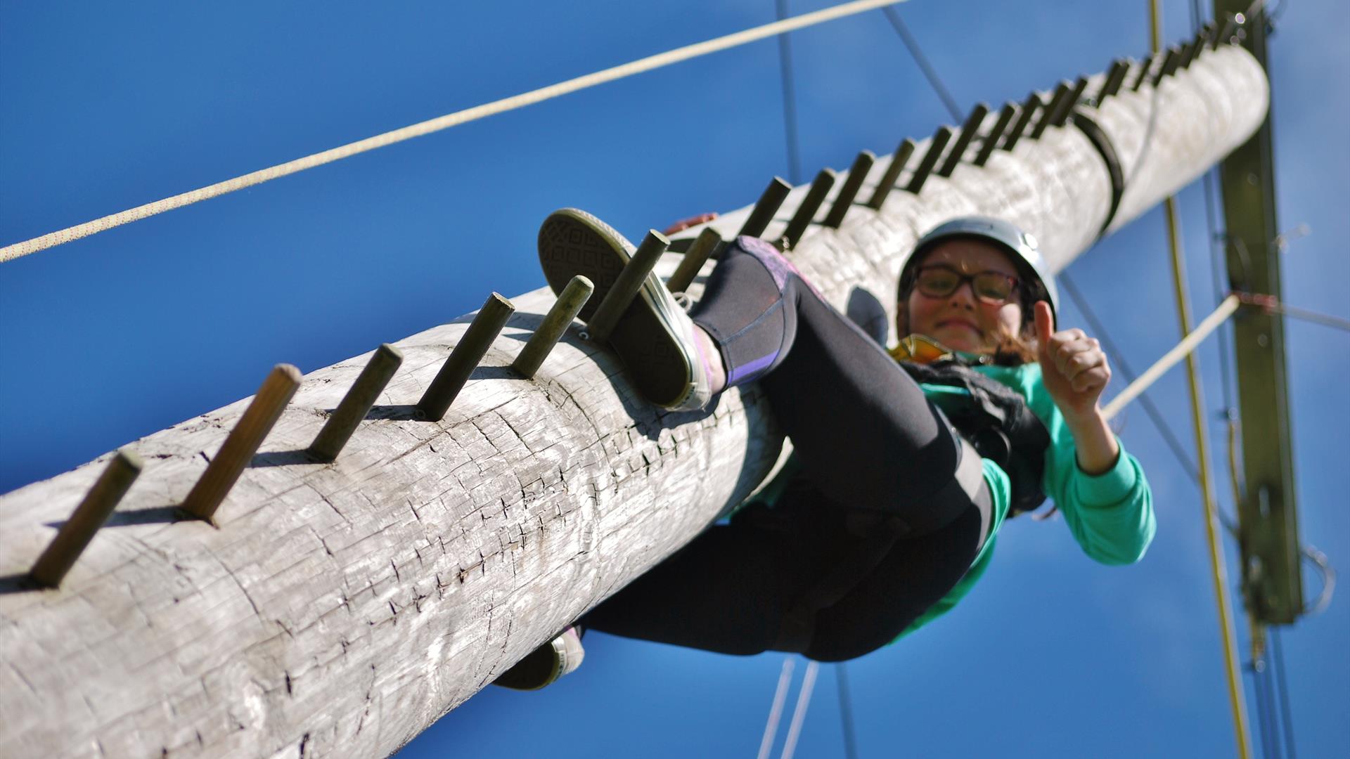 A young girl climbing up a wooden climbing pole wearing protective clothing
