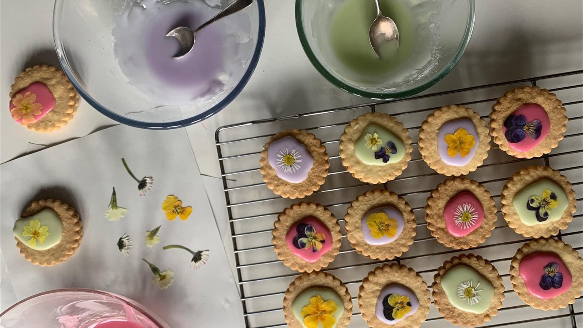 Image shows bowls of pastel coloured icing and some biscuits decorated with the iciing and with little edible flowers.