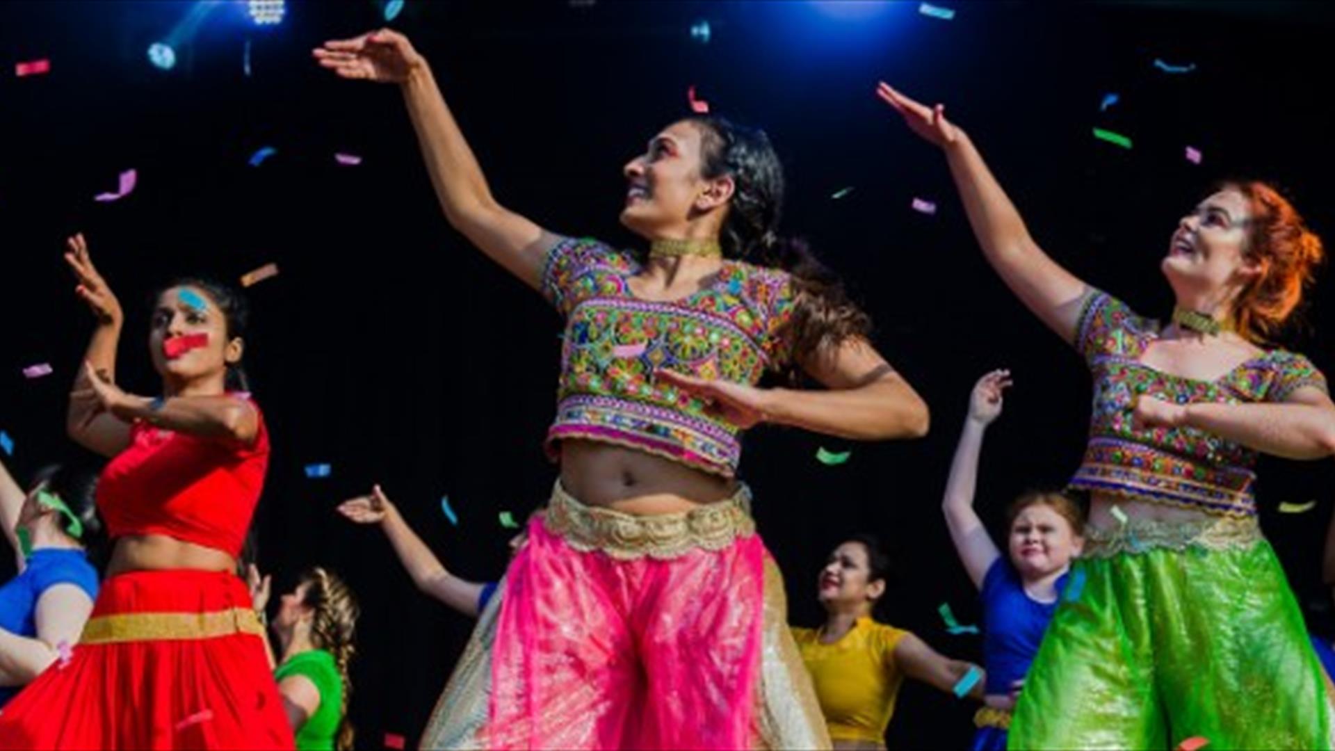 Image shows three female dancers wearing brightly coloured clothes and with one hand up in the air