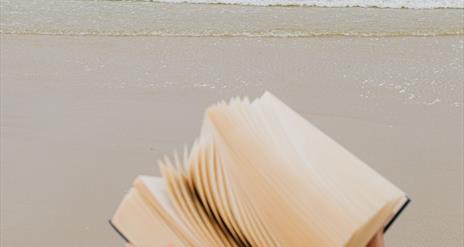 Close up image of hands holding a book with the sea and sand behind them,.