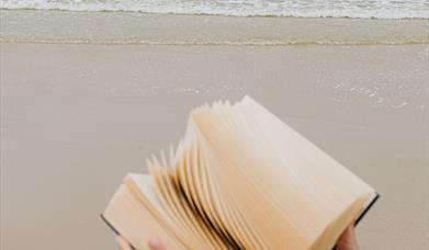 close up of hands holding a book with the sand and the sea behind them.