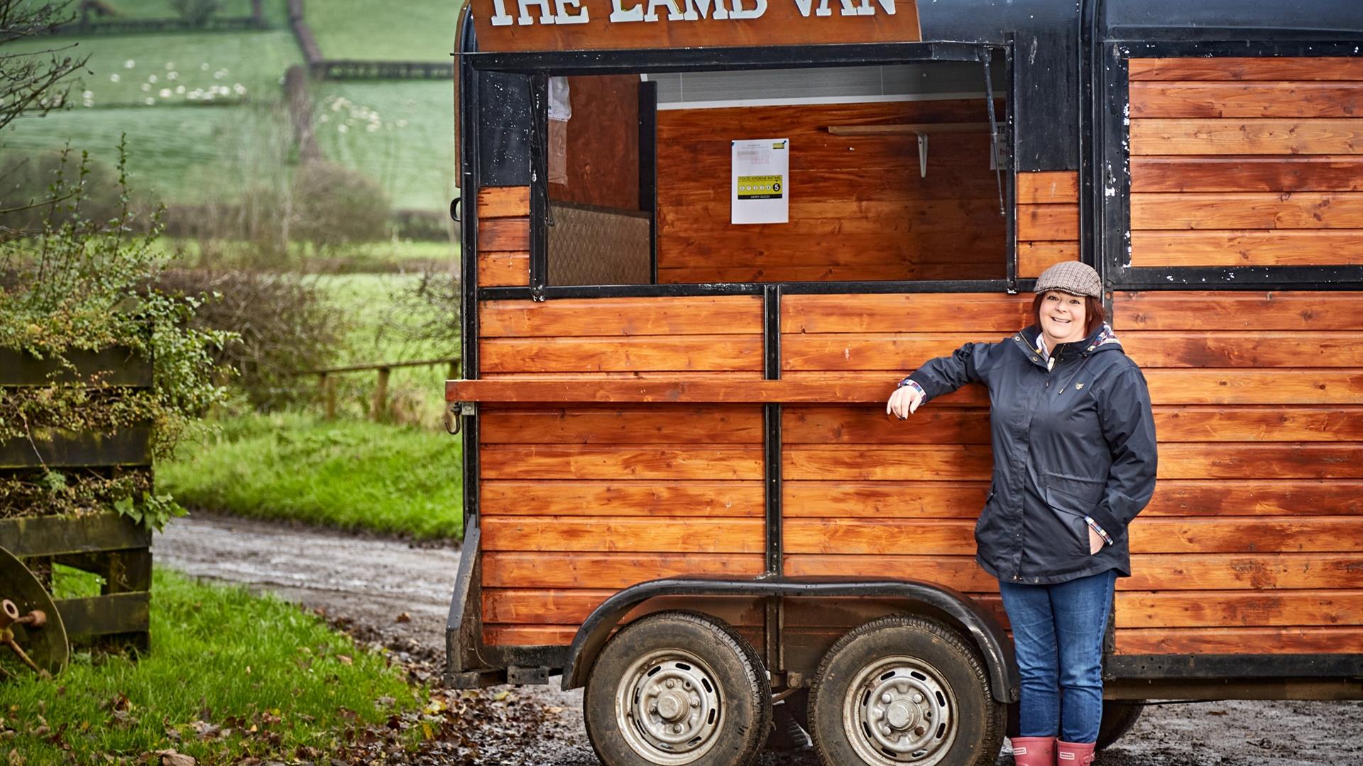a woman wearing a flat cap and wellies stands beside a wooden food truck which has a sign that reads 'The Lamb Van' - countryside fields are visible i a woman wearing a flat cap and wellies stands beside a wooden food truck which has a sign that reads 'The Lamb Van' - countryside fields are visible i