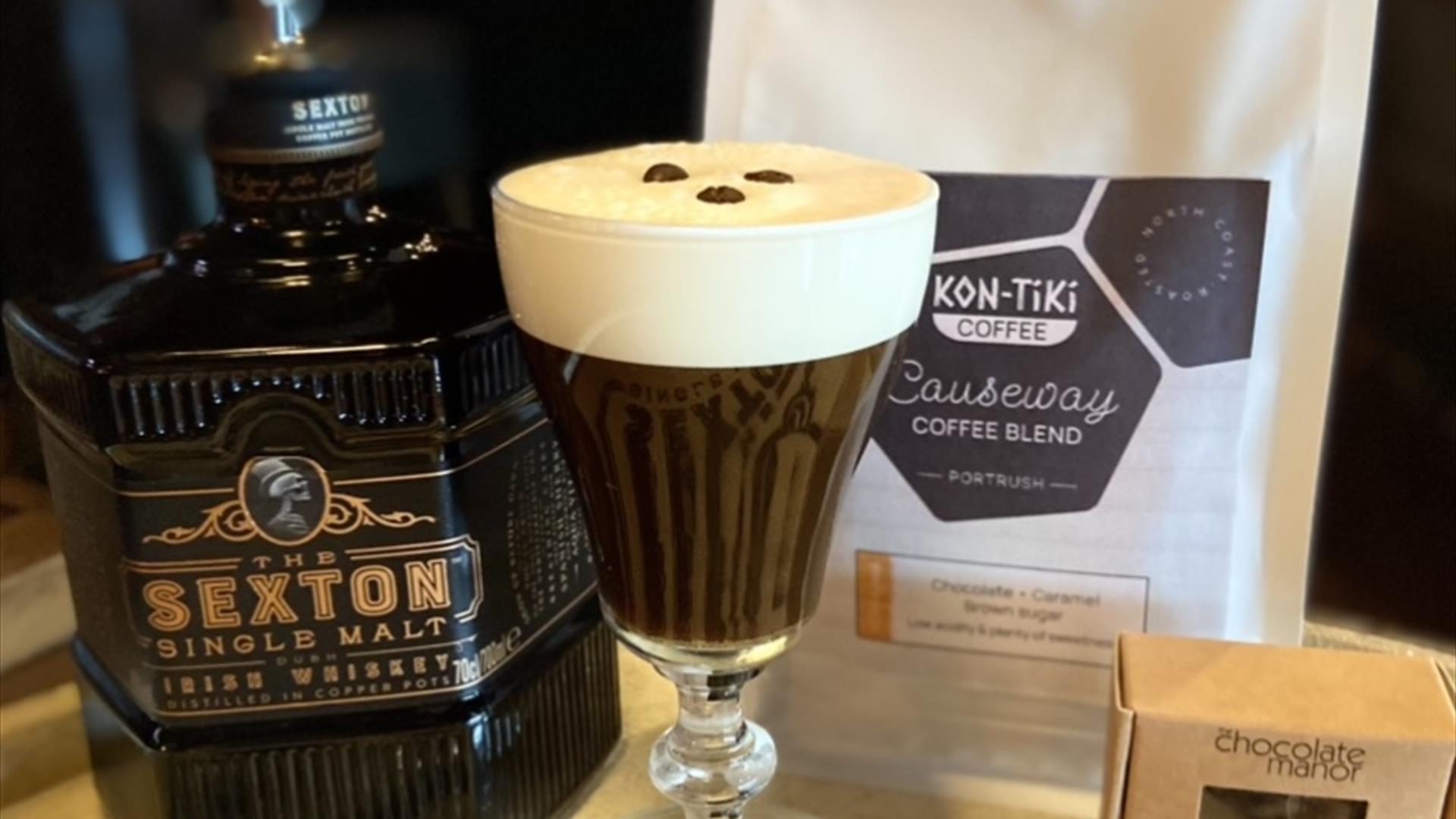A still life of a bottle of sexton single malt whiskey, an Irish Coffee with 3 coffee beans floating on the cream head, a pack of Kon-Tiki Causeway Co