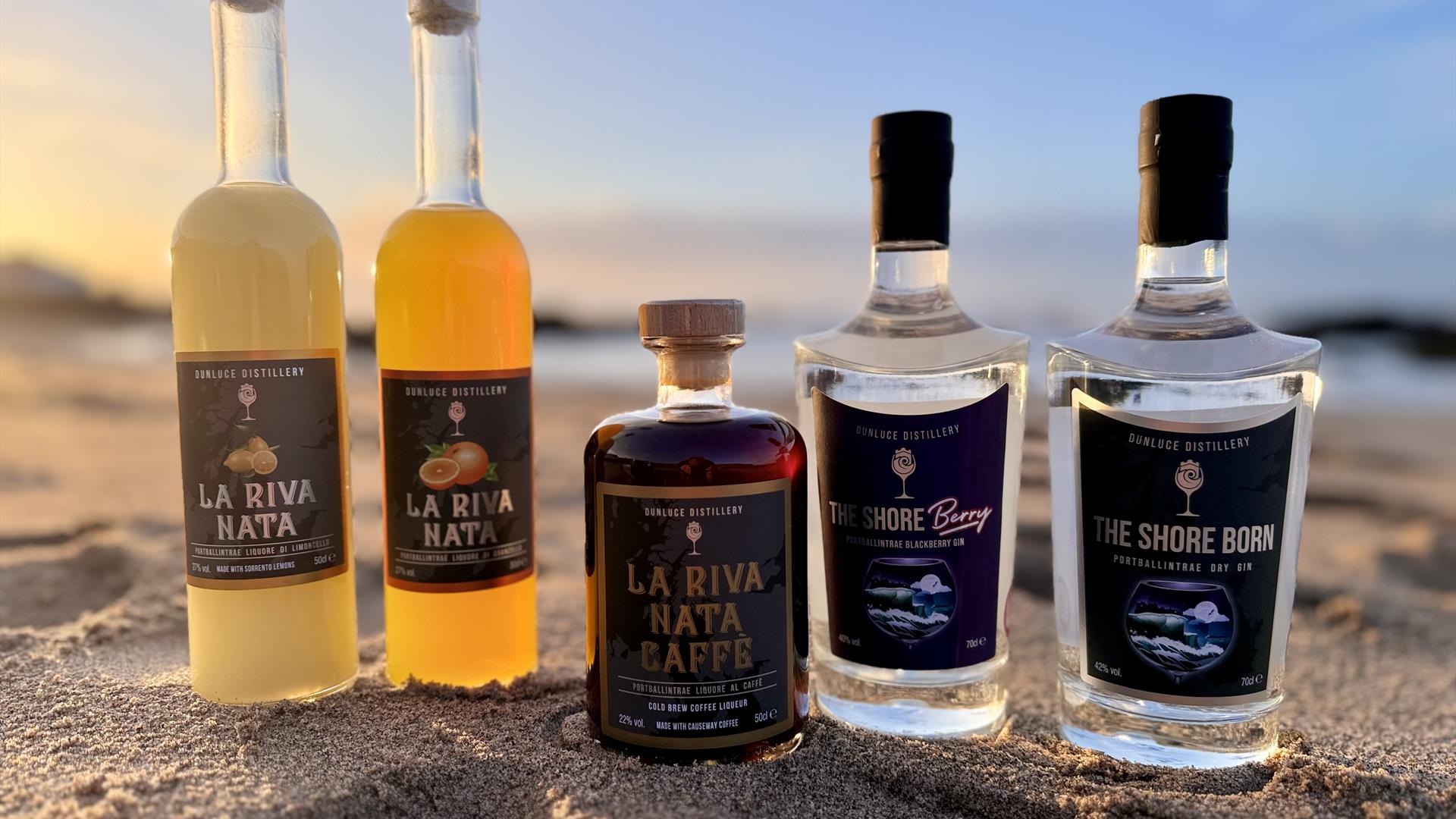 various bottles of alcohol including gins and liqueurs produced by Dunluce Distillery are lined up together on the sand at the beach various bottles of alcohol including gins and liqueurs produced by Dunluce Distillery are lined up together on the sand at the beach