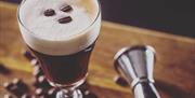 Image taken from above of an Irish Coffee surrounded by a scattering of coffee beans on a wooden bench with a metal measure lying to the right of the