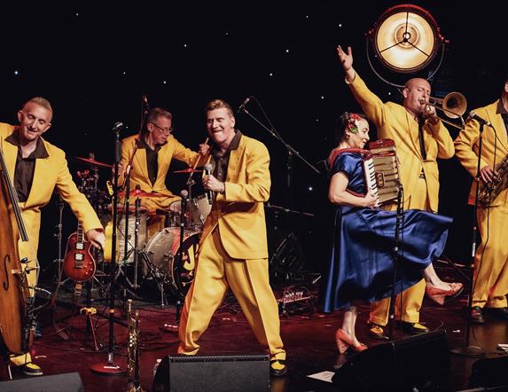 Image shows a band on stage with men in yellow suits and a women in a blue dress