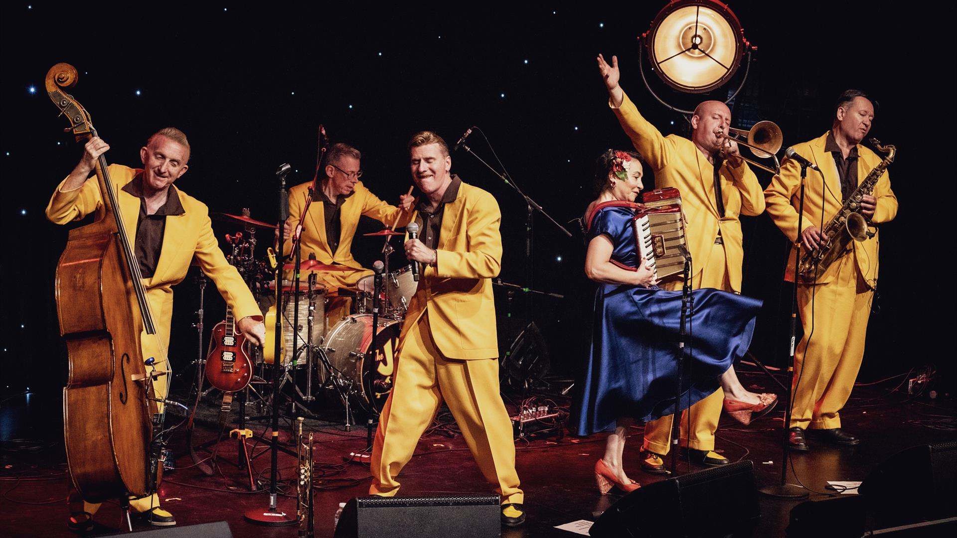 Image shows a band on stage with men in yellow suits and a women in a blue dress
