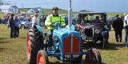 Image of a gentleman driving a blue vintage tractor. He is wearing a high visibility vest, a flat cap and is smoking a pipe.  Just behind him is a mar