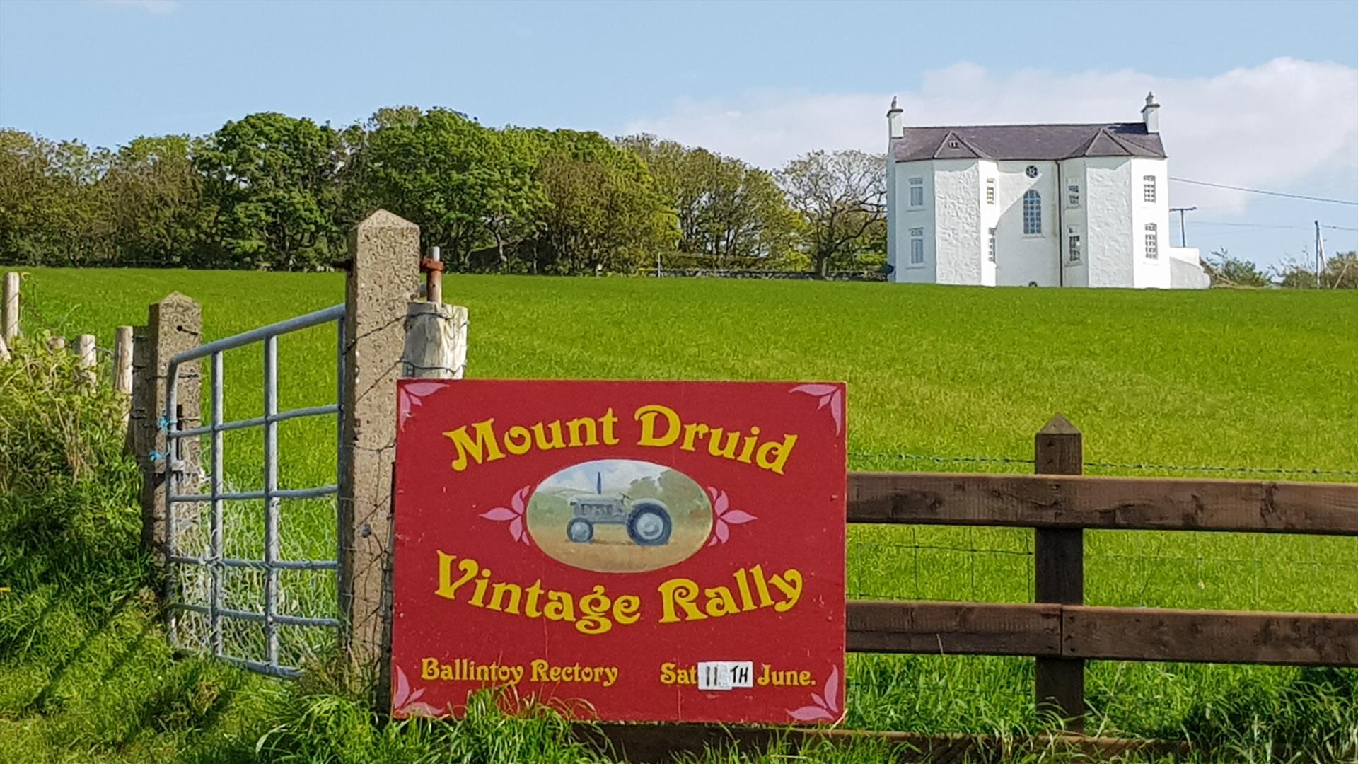 Image showing Ballintoy Rectory in the background with green fields in the foreground.  There is a sign on a fence advertising the Mount Druid Vintage