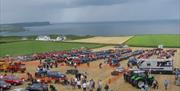 Aerial image of the rally, showing green fields leading out to the sea with tractors, caravans, cars and people milling about looking at the vehicles