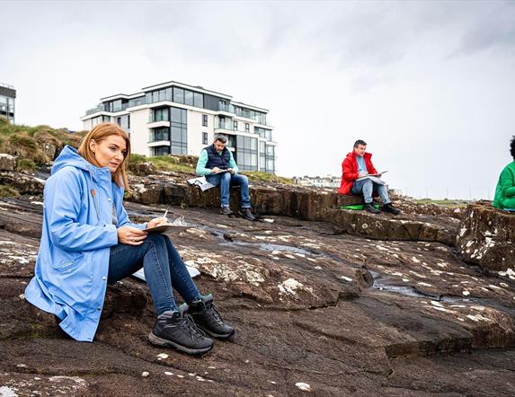 Participants of the workshop sit with sketch pads on coastal rocks in Portrush