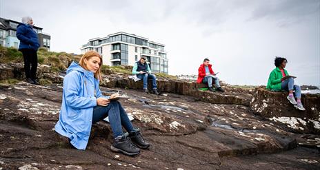 Participants of the workshop sit with sketch pads on coastal rocks in Portrush