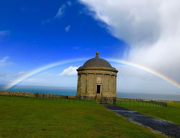 Image of the Mussenden Temple surrounded by green grass and a blue sky, clouds and a rainbow above