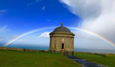 Image of the Mussenden Temple surrounded by green grass and a blue sky, clouds and a rainbow above