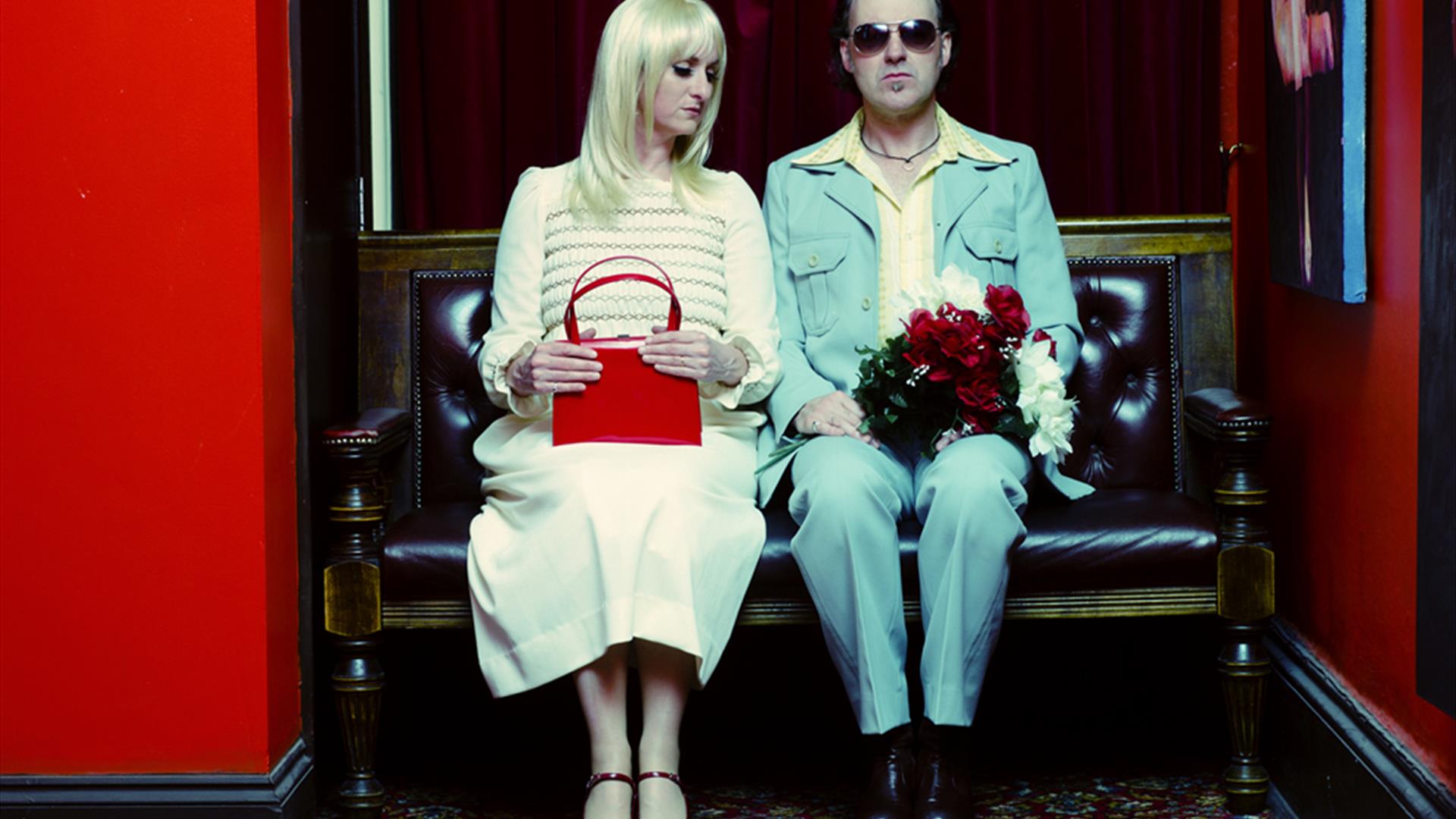 a man and woman in vintage clothing sitting on a bench. The woman is holding a red handbag, the man is holding a bouquet of red and white flowers.