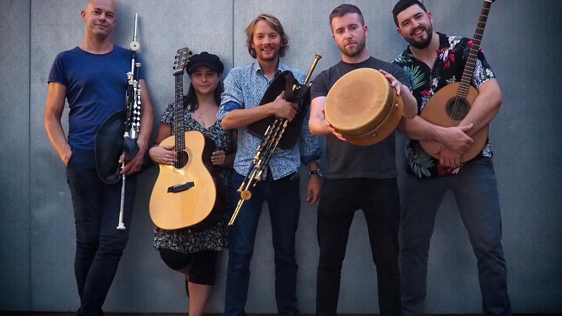 Image shows Realta Band standing outside.  5 members of the band 4 male and 1 female stand with instruments such as guitars and bodhran.