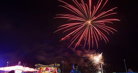 Image shows fireworks lighting up the night sky with the crowd and fairground amusements below