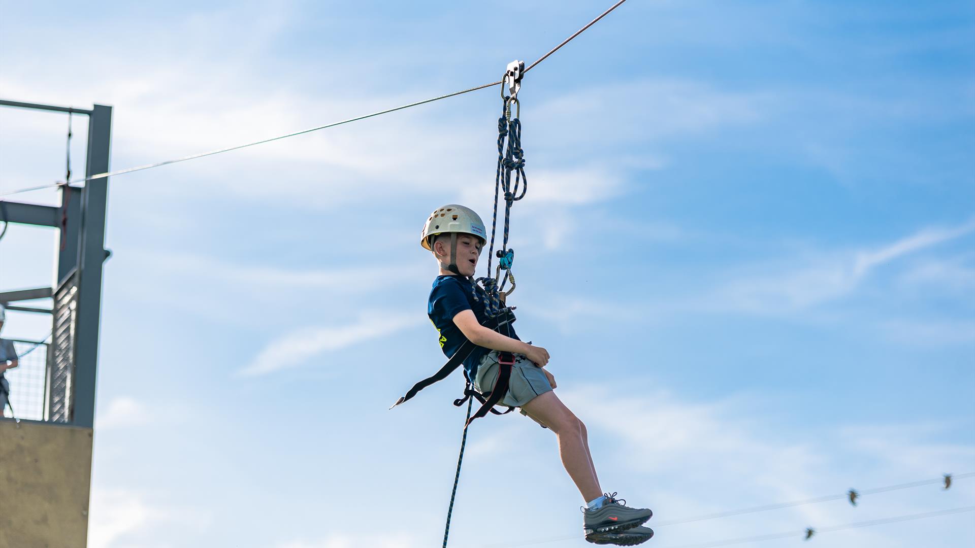 A boy in a blue outfit and white helmet harnessed to a zipline