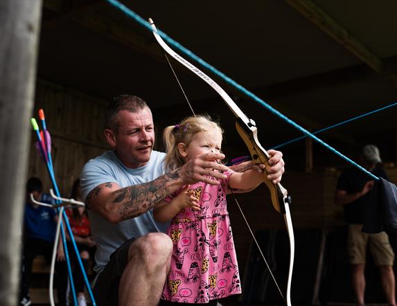A man in a blue t-shirt and shorts helping a little girl in a pink dress with a bow and arrow
