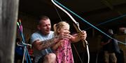 A man in a blue t-shirt and shorts helping a little girl in a pink dress with a bow and arrow