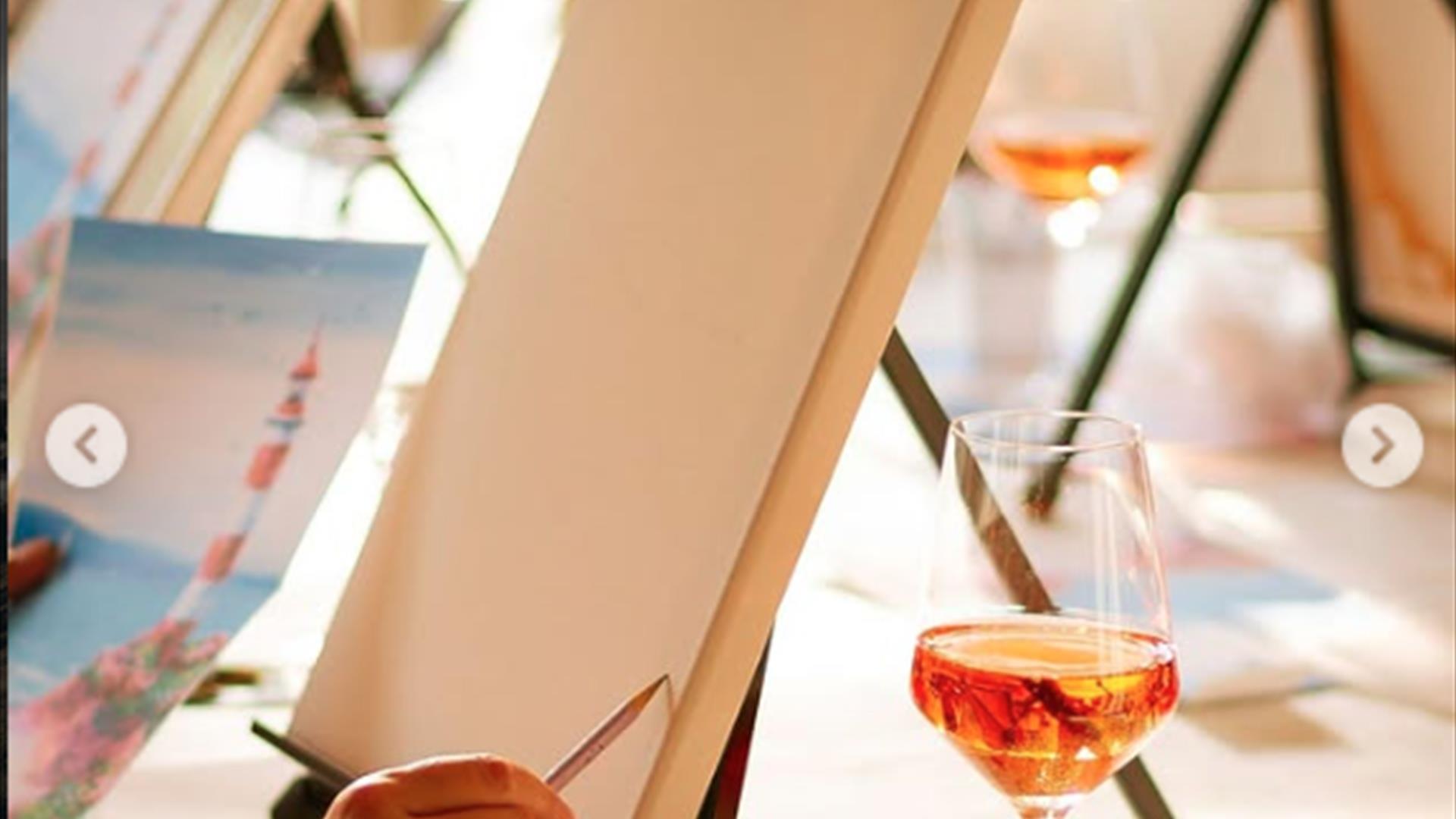 a person holds a pencil against a blank canvas resting on an easel. There is a glass of wine and a small plastic cup sitting on the table beside it. a person holds a pencil against a blank canvas resting on an easel. There is a glass of wine and a small plastic cup sitting on the table beside it.