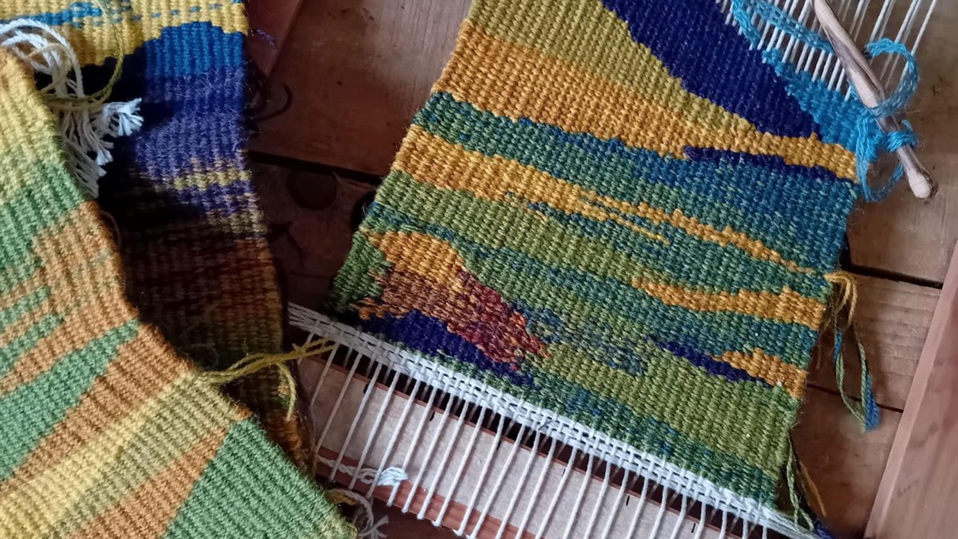 three colourful tapestries - one placed in a loom - sit on a wooden surface three colourful tapestries - one placed in a loom - sit on a wooden surface
