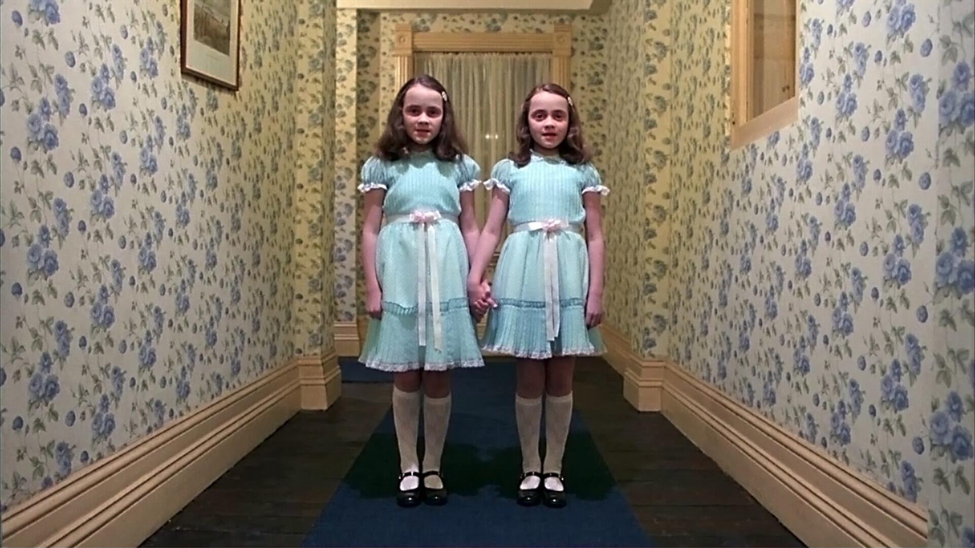 Twin girls holding hands in the hallway - both wearing the same blue dress with a white ribbon around the waist