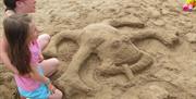 Image shows a large sand sculpture of a frog with two young girls who made it sitting beside it.