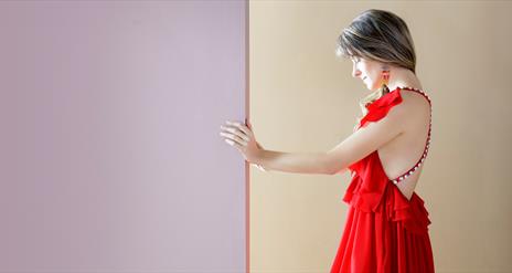 Image shows a woman wearing a red dress holding her hand against a wall. The left side of the wall is purple and the right side cream.