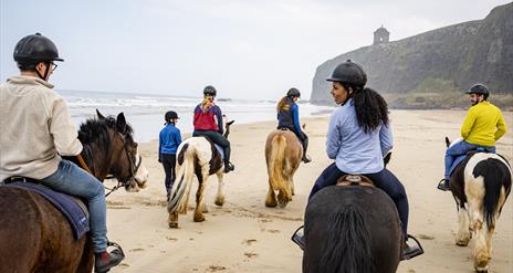 A group of people on horses riding down the beach with the Mussenden Temple in the background