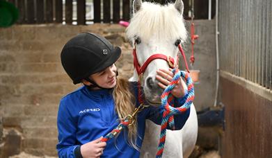 Image shows a young girl in a blue top and black riding helmet petting a white horse