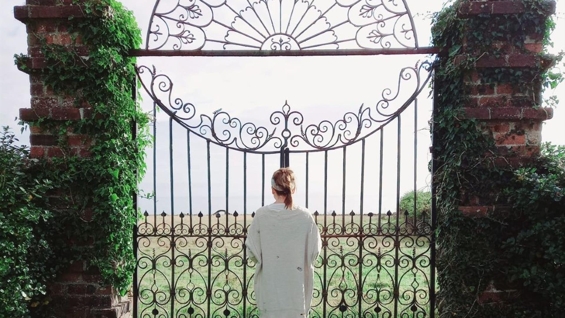 Person facing wrought iron gate with fields in background