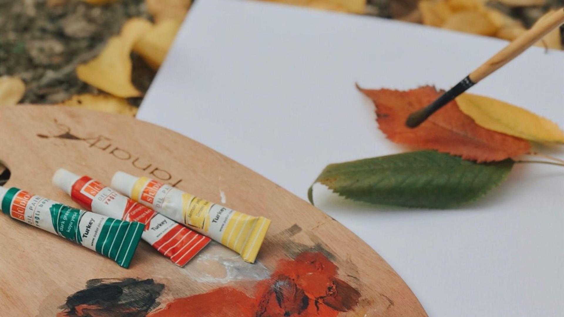 an artist's palette holding tubes of green, orange and yellow paint and a paintbrush sits on the forest floor beside a blank page which has leaves on an artist's palette holding tubes of green, orange and yellow paint and a paintbrush sits on the forest floor beside a blank page which has leaves on