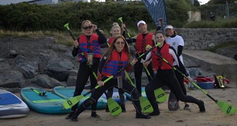 a group of people posing with paddles on a beach with paddleboard in the background