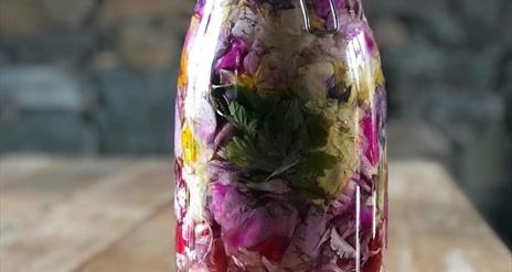 Image shows a close up of a jar filled with liquid and colourful herbs.